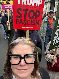 A close up of me, with flat, windswept hair. Behind me are other protesters, lots of people with signs and flags. The main sign that's front and centre behind my head says, Stop Trump, Stop Fascism.''