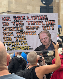 A loser of a sign held up by a protester. It has a picture of Biff from Back to the Future, and the wording says, 'We are living in the timeline where Biff got his hands on the sports almanac.'