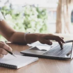 Individual multitasking by writing in a notebook and using a laptop.