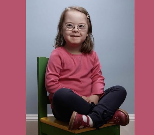 young Penny is wearing a pink long-sleeve shirt, dark leggings, and red shoes and sits cross-legged on a small green-and-yellow chair against a light blue background. She is smiling slightly and has a hair clip on one side.
