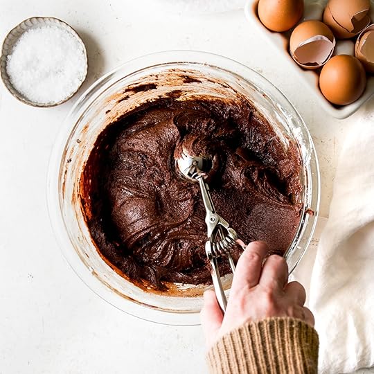 Overhead shot of rich, fudgy chocolate cookie dough being scooped from a clear glass bowl with a metal scoop, alongside a bowl of flaky sea salt and cracked eggs for the easy peanut butter cup cookies recipe.