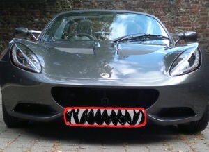 Picture showing a grey, low-slung, fast looking sports car against a flint and brick wall. The numberplate has been blacked out in the picture, so as not to show the real one on t'interweb, and the photographer has put a red line round the outside of the hole where the numberplate should be shown (as if it's a pair of lips) and drawn in teeth.