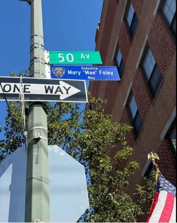 A street pole with green '50 AV', blue 'Detective Mary Mae Foley Way', and a black-and-white 'ONE WAY' sign, near red brick, trees, and flag.