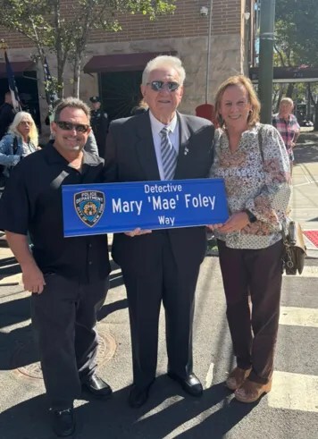Three adults stand outdoors, smiling, holding a street sign that reads 'Detective Mary Mae Foley Way.' Each wears different outfits.