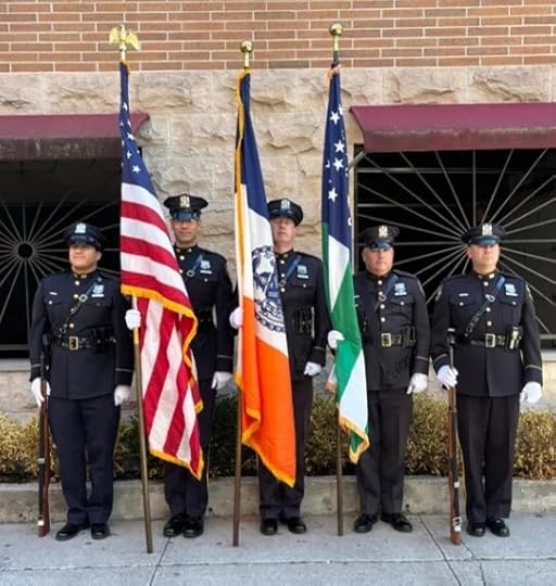 Five police officers in uniform hold three flags—U.S., NYC, and one with a green stripe—outside a stone and brick building.
