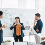 A group of professionals discussing a project, with tasks outlined on a whiteboard during a meeting.