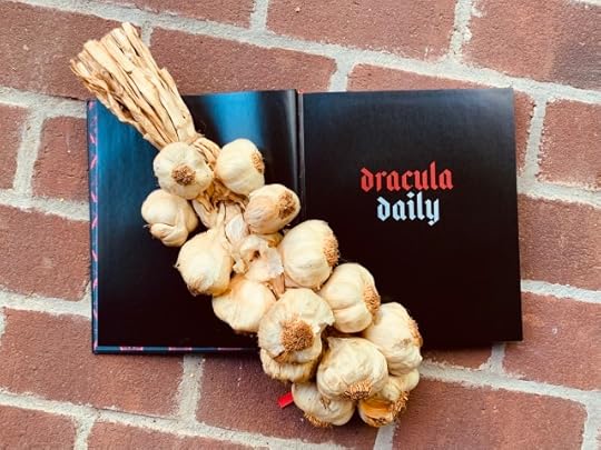 Pictured is the book Dracula Daily resting on a brick walk with a bunch of garlic resting on its open pages.