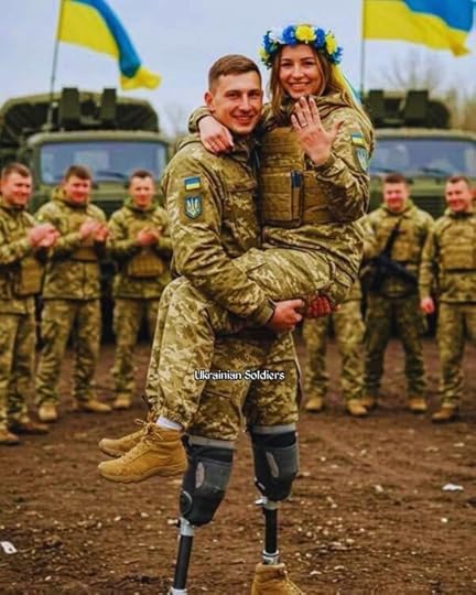 Two Ukrainian soldiers in camouflage uniforms, one carrying the other, both wearing floral crowns. They stand on a dirt path with military vehicles and other soldiers in similar uniforms clapping in the background. Ukrainian flags are visible, and a watermark reads 