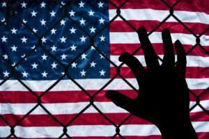 A hand grabs at a chain link fence with an American flag on the other side, symbolizing the struggle to immigrate to America.