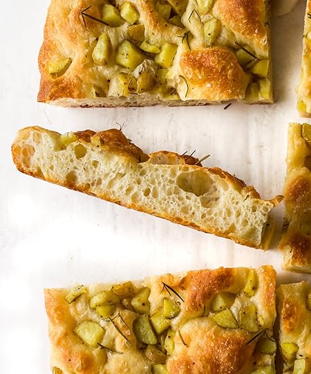 A close-up shot of several sites of herby potato focaccia with one long side turned on its side to clearly show the airy, bubbly cross-section (crumb) of the bread, which is golden brown on top and features pieces of potato, herbs, lemon zest, olive oil and sea salt flakes.