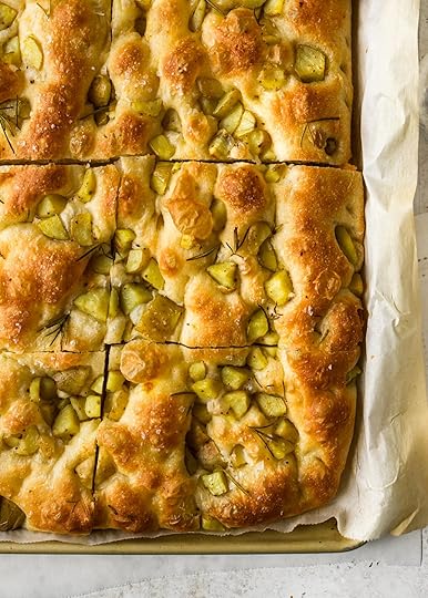 An overhead close-up shot of a large, golden brown sheet of herby potato focaccia, freshly baked in a rectangular pan lined with parchment paper, topped with small potato pieces, fresh rosemary sprigs and flaky sea salt, cut into slices.