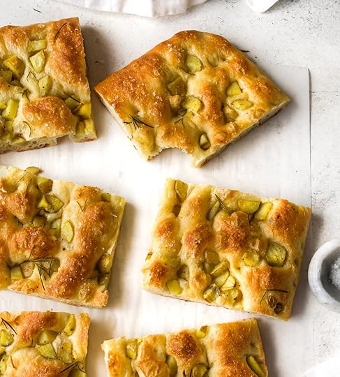 An overhead arrangement of several individual squares of golden-brown herby potato focaccia, each piece dimpled and topped with chopped potatoes, fresh rosemary and flaky sea salt, scattered across white parchment paper next to a small marble salt cellar.