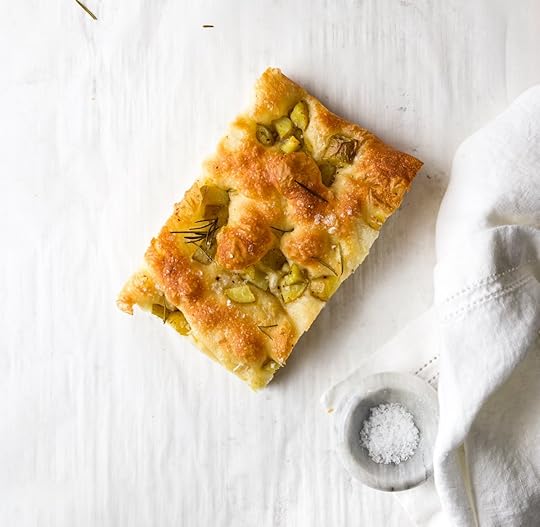 A square slice of golden brown, herby potato focaccia, dimpled on top and sprinkled with flaky sea salt, resting on white parchment paper next to a small marble bowl of salt and a white cotton napkin.