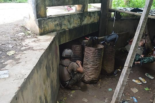 Soldier taking cover behind barrels in a tactical environment, showcasing military readiness and camouflage in a jungle setting.