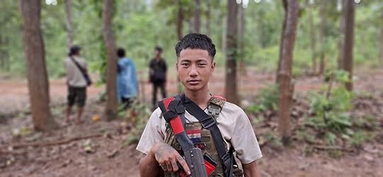 Young soldier in military attire holding a firearm in a forested area, with other individuals visible in the background.