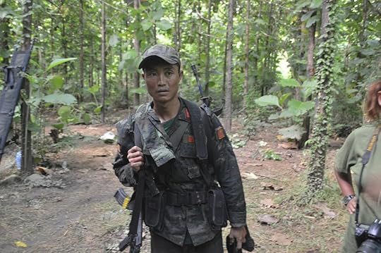 Soldier in military attire stands in a dense forest, holding a weapon, with foliage and trees surrounding him.