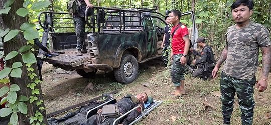 Military personnel gather near a pickup truck in a forested area, examining a fallen comrade on a stretcher.