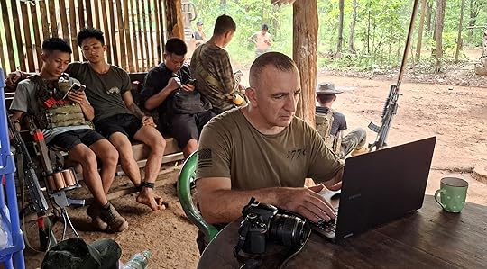 A military personnel works on a laptop at a makeshift camp while others relax nearby, surrounded by nature.