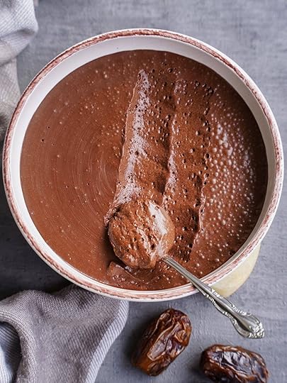 A bowl of chocolate mousse with a spoonful taken out, placed on a gray surface with two dates nearby