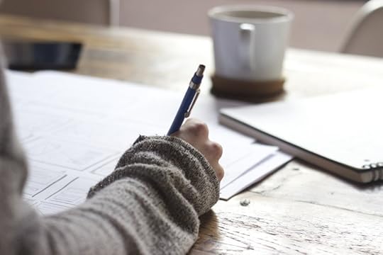 Public-domain image of a hand holding a pen, apparently writing, at a sunlit desk with papers and a white coffee mug on it. The sleeve of a cozy gray sweater is visible.