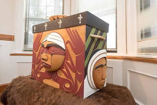 A Bentwood Box by artist Luke Marston, featuring intricate carvings representing Indigenous heritage, displayed indoors near a window.