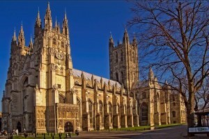 A photograph of Canterbury Cathedral in Canterbury, England, which is a medieval cathedral made from light colored stone, with flying buttresses, towers, and lots of exterior architectural ornament.