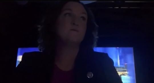 Person speaking in a dimly lit setting with a blue background, wearing a black jacket and looking thoughtful.