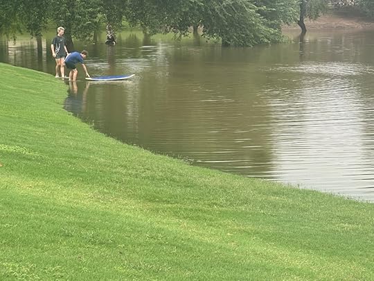 Two individuals observed on the green grass by a flooded area, one preparing to balance on a paddleboard while the other watches.