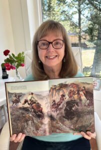 Smiling woman with brown hair and glasses holds open a book with a colourful illustration by Charles Russell of cowboys roping horses.