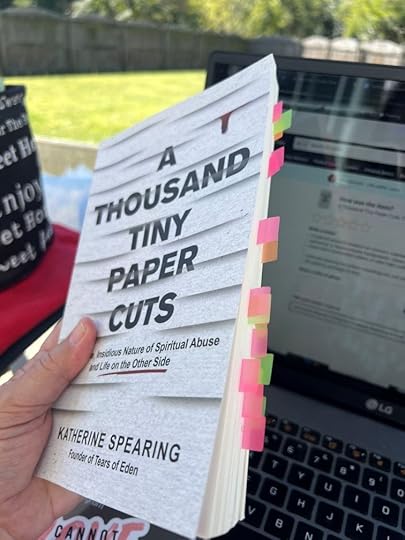 A hand holds a copy of the book A Thousand Tiny Papercuts: The Subtle, Insidious Nature of Spiritual Abuse and Life on the Other Side by Katherine Spearing. The book has multiple colorful sticky tabs marking pages. In the background, there’s an open laptop and a green backyard visible in bright sunlight.