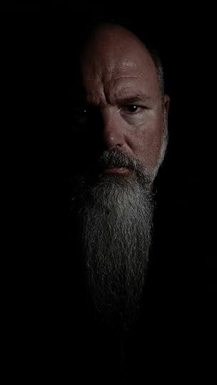 a low-key portrait showing half a white man's face emerging from darkness. The image features a bald man with a long gray beard, with strong side lighting creating contrast that emphasizes an intense expression and facial features while leaving the rest in shadow.