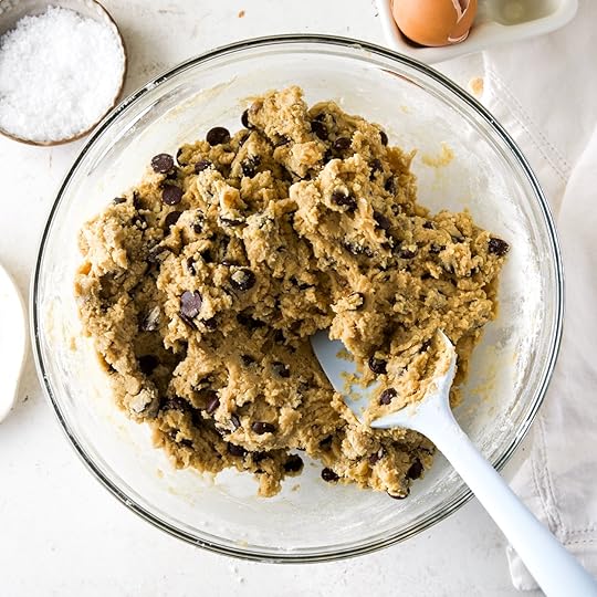 A bowl of delicious chocolate chip cookie dough made with walnuts in a glass bowl with a light blue spatula sticking in it.