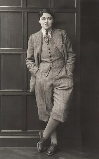 A 1920s woman with slicked down short hair with a sharp side parting, wearing eyebrow and eye makeup, stands confidently against a wooden wall, dressed in a vintage gray suit with knickerbockers, a tie, and dark shoes, exuding a stylish and sophisticated demeanor.