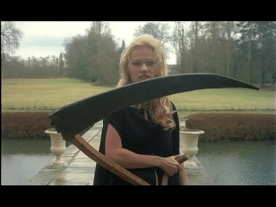 A woman with long blonde hair, dressed in a long black sleeveless garment, stands on a stone path over a moat, holding a large scythe, with a grassy area and trees in the background.
