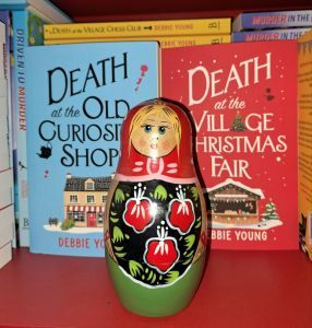 Paperback copies of Death at the Old Curiosity Shop and Death at the Village Christmas Fair with a traditional Russian doll