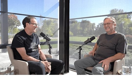Two men sitting in chairs in a bright, modern setting, engaged in conversation with microphones in front of them.