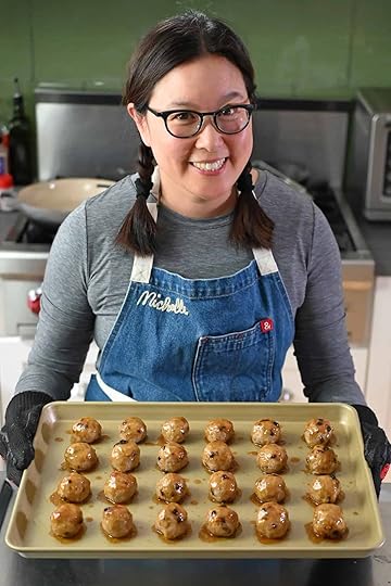 A smiling Asian woman in glasses is holding a gold rimmed baking sheet topped with freshly baked cranberry turkey meatballs topped with a maple dijon mustard glaze.