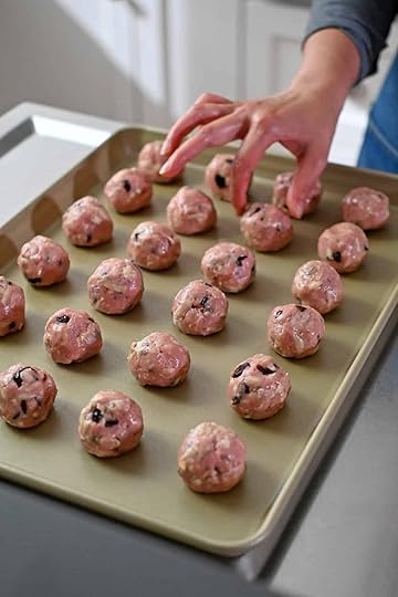 A hand is placing raw turkey meatballs with cranberries on a gold rimmed baking sheet.