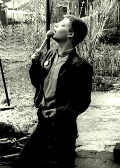 Sepia-toned black and white image of a short-haired white woman wearing wire frame glasses and a leather jacket singing into a microphone somewhere outdoors