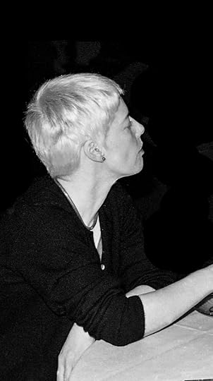 Short, platinum-haired white woman sitting at a table watching something off camera