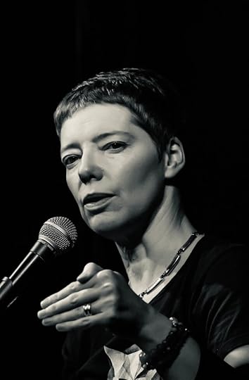 BW image short-haired white woman with eyes narrowed in intensity as she gestures and talks into a microphone before an off-camera audience