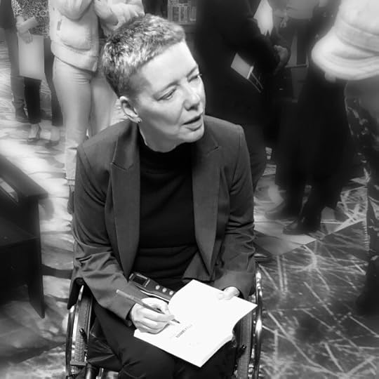 Short-haired white woman sitting in a wheelchair. She is wearing a black turtleneck and grey suit jacket and signing a book