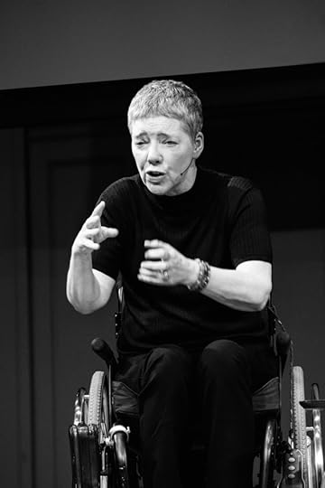 Short-haire white woman wearing headset mic leaning forward intently in erh wheelchair and gesturing as she talks to the large audience below the stage