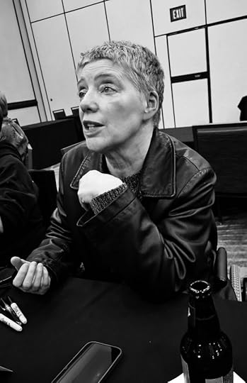 Short-haired white woman sitting in a wheelchair behind a table looking up and talking to someone off camera