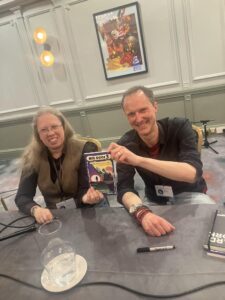 Two authors - Ruthanna Emrys and Andrew Knighton - sit behind a table grinning and hold their book.