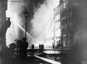 A black and white photograph of firefighters trying to extinguish burning buildings at night during the London blitz.