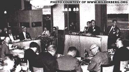 Historical image of the first Bilderberg Meeting in 1954, showing attendees seated at tables with a speaker at a podium, discussing key geopolitical issues.