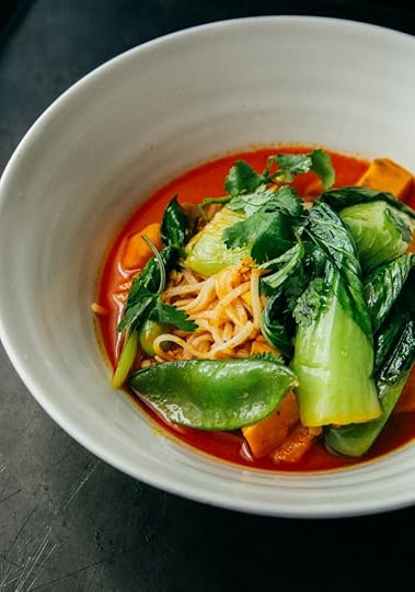 A bowl of noodle soup with vibrant orange broth, topped with fresh greens such as bok choy and cilantro, along with other vegetables.