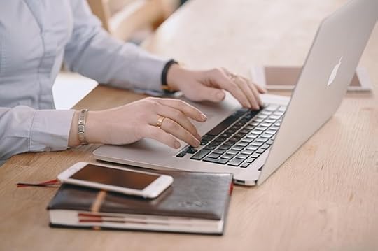 On a table sit an open laptop, a leather-bound book with a smartphone on top, and a tablet in the background. A woman's hands are typing on the laptop's keyboard.