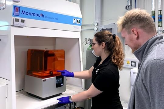 A large chamber surrounds a desktop resin printer as a gloved instructor explains how to use it.